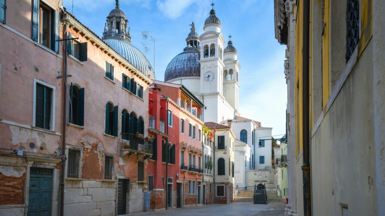 View of the domes and bell tower at Basilica di Santa Maria della Salute on the Grand Canal in Venice. Narrow streets and typical architectural style of Venice, Italy. Old houses along the canal.