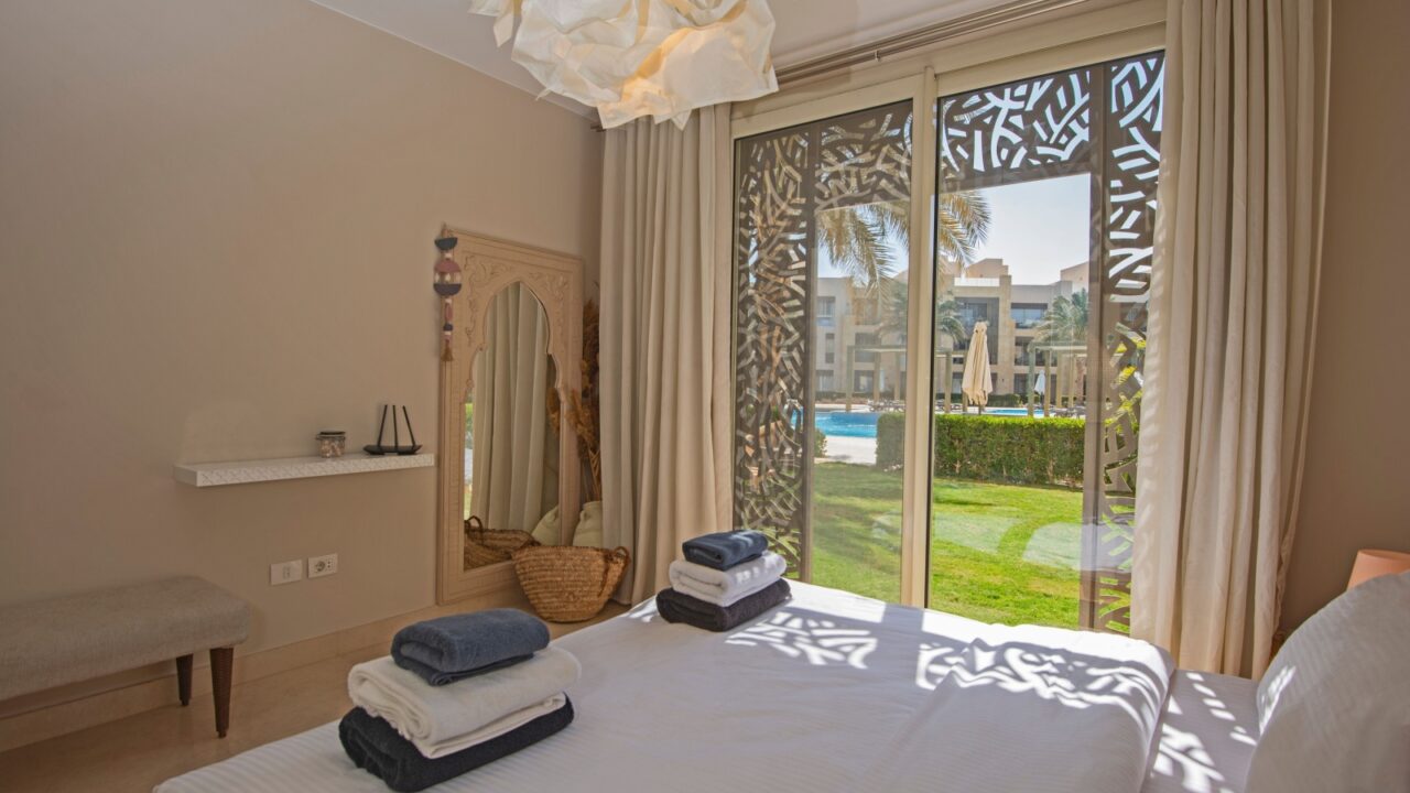 Bedroom with white linens, nautical decor, patterned screens, garden view, pool, palm trees, and sunlight.