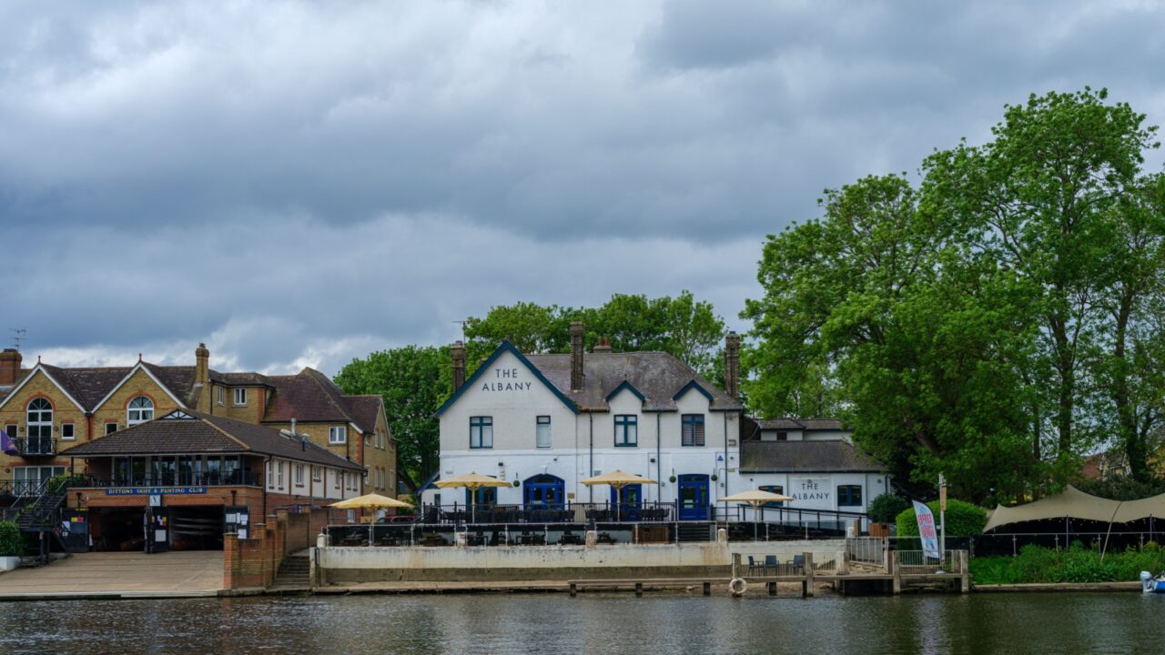 UK, Hampton Court, 05 May 2025 Riverside buildings with outdoor terraces face the calm Thames waters near Hampton Court, offering scenic views and relaxed dining by the river.