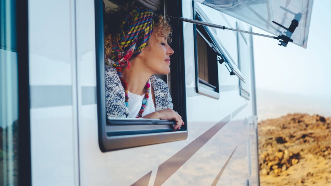 Camper in a desert setting with a curly-haired woman with a scarf on her head leaning against the window of the camper. van with tourist stop on the mountain side to get a better view of the view.