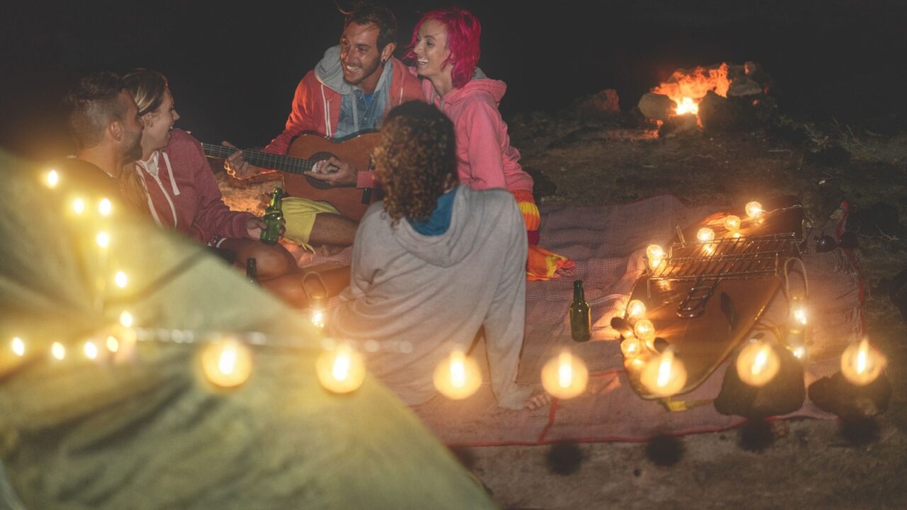 Group of friends having fun playing guitar and laughing together in desert next sea at night time - Young people camping during summer holidays - Travel and friendship concept - Focus on top man face