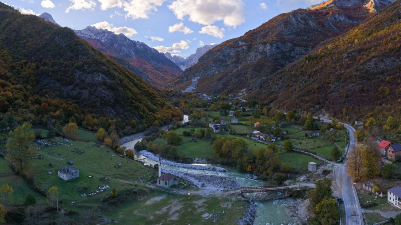 Aerial panorama of Dragobi village in Valbona Valley National Park with the turquoise Valbona River flowing between forested mountains.