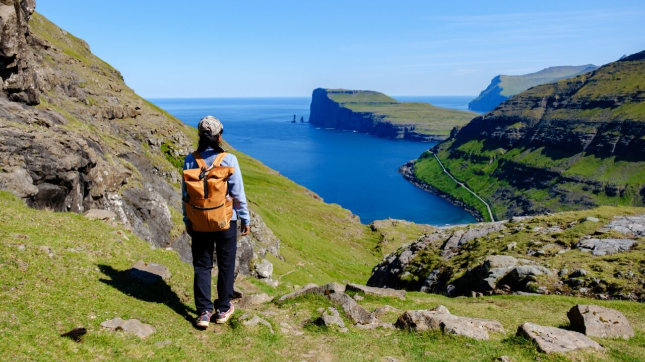 A traveler gazes at the breathtaking cliffs and fjords of the Faroe Islands on a sunny summer day. The vibrant green hills lead to the sparkling blue waters, showcasing natures grandeur.