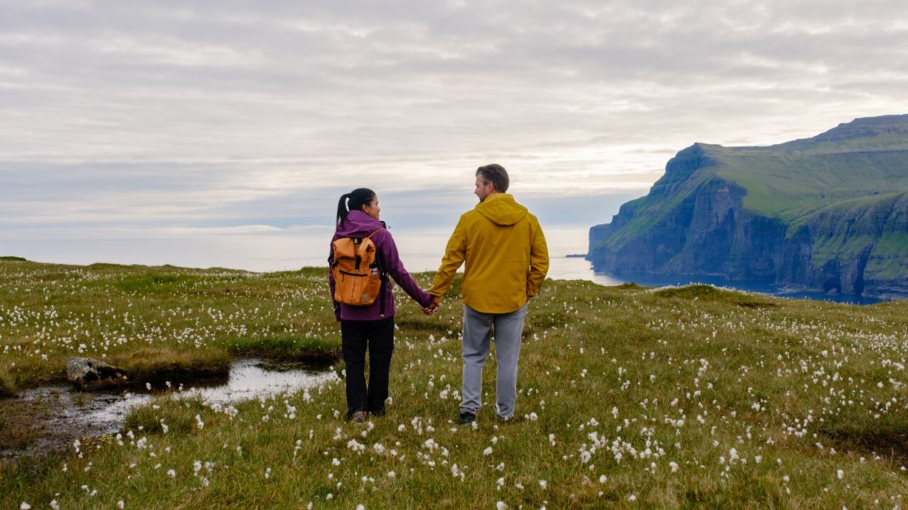 A couple walks hand in hand across a vibrant meadow filled with cotton grass in the Faroe Islands, surrounded by stunning cliffs and tranquil waters at dusk. Eidiskollur Eidi mountain