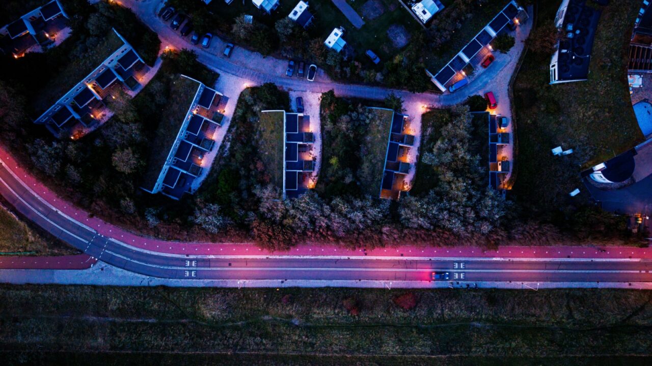 Aerial view of twilight residential zone curved housing grid, lit multi lane road with red bike paths, and grassy field below blend geometric planning with natural contrast.