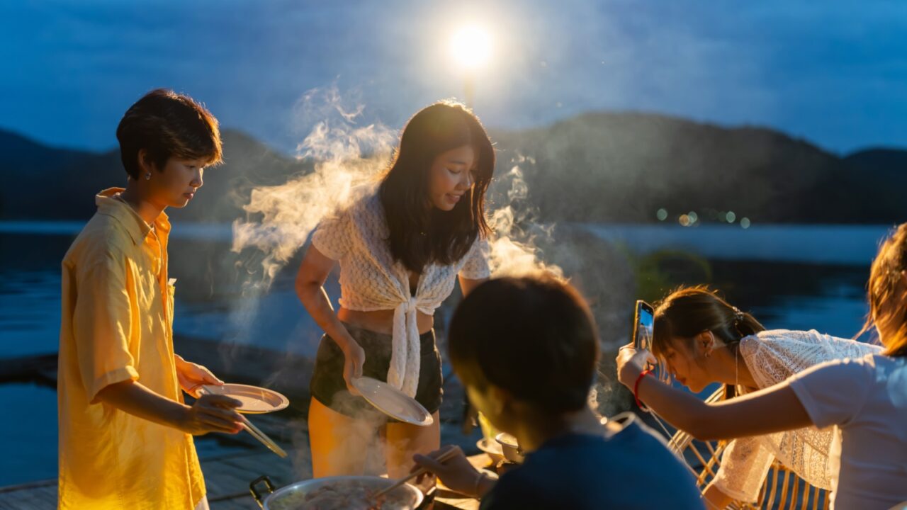 Group of Young Asian man and woman friends enjoy and fun celebration meeting dinner party eating barbecue grill and drinking beer together on lake house balcony on summer holiday vacation at night.