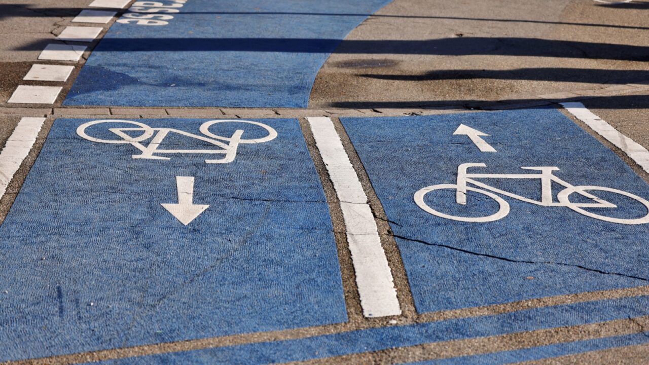 Urban bicycle lane with blue pavement and white directional bike symbols. Divided for two-way cycling traffic, marked with arrows and dashed lines. Promotes safe and organized cycling infrastructure.
