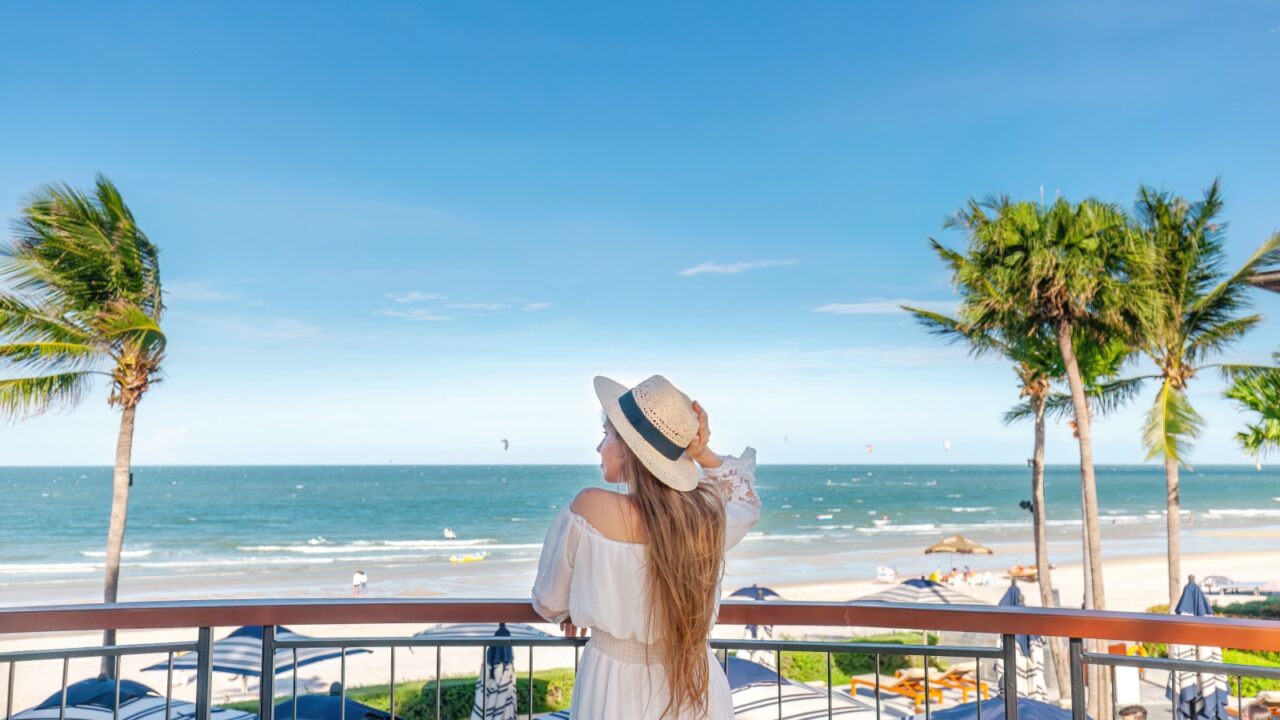 Sea view from balcony with woman in hat and dress, overlooking beach activities and kitesurfers on water. Tropical vacation ambiance.