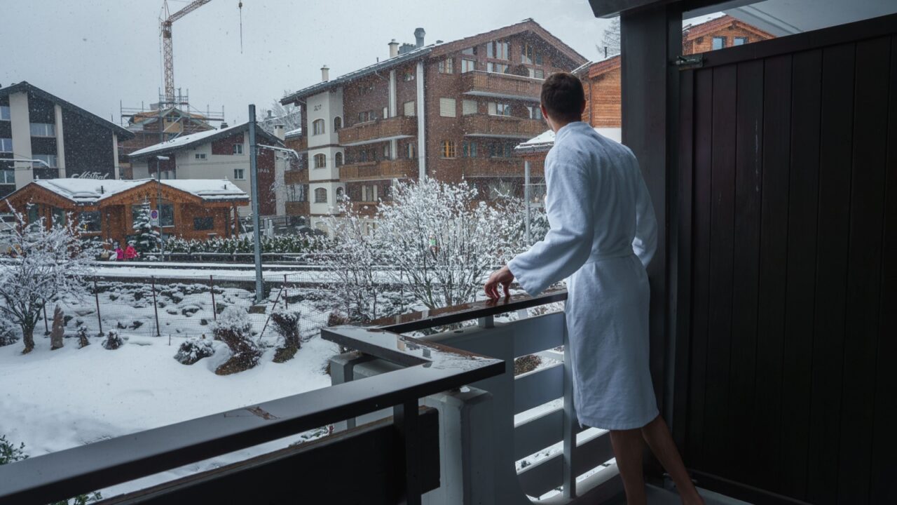 Luxury hotel balcony view in Zermatt, Swiss ski resort. Person in bathrobe gazes at snowy landscape. Alpine architecture and ongoing development visible. Snowfall adds tranquil atmosphere.