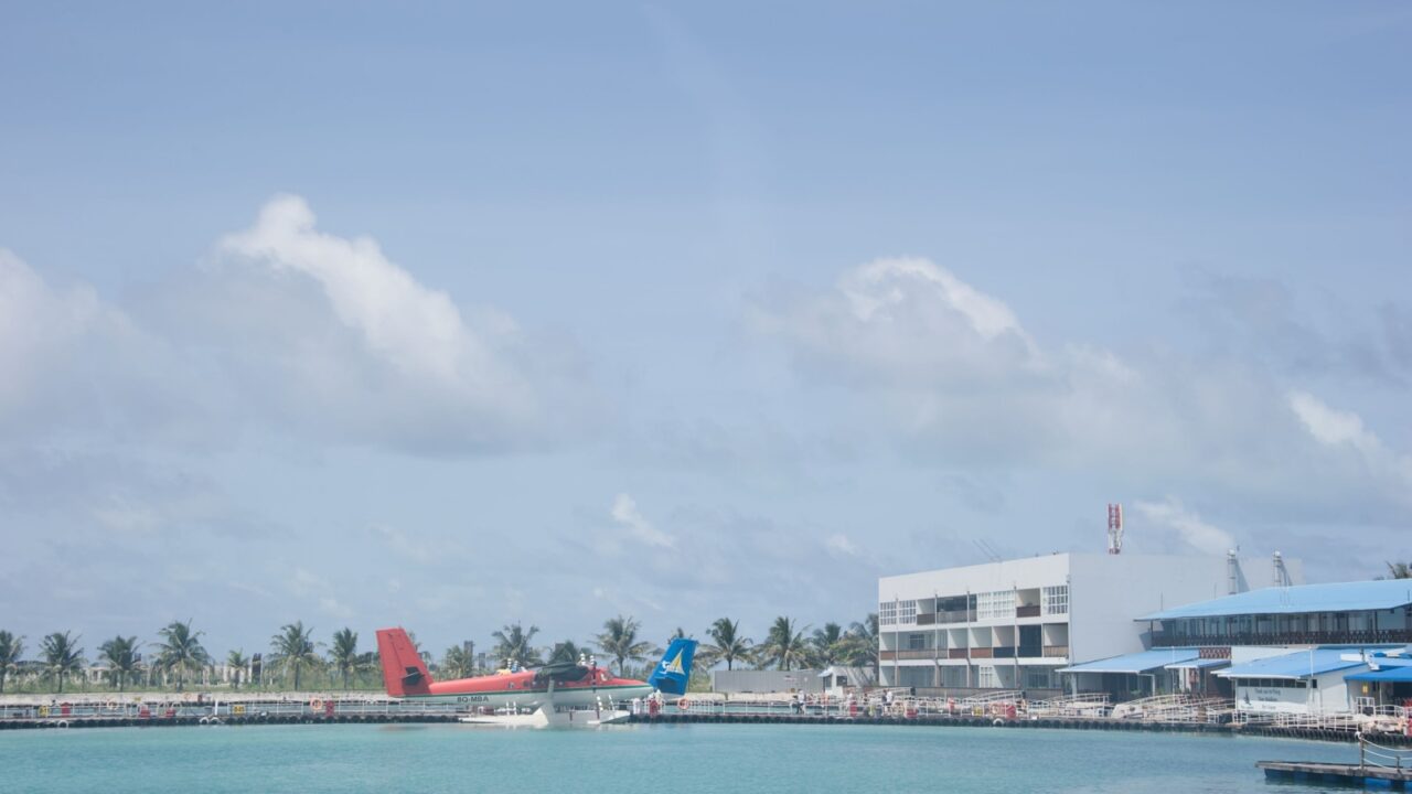 Velana International Airport (MLE), Aug 12, 2014: Wide view of the seaplane terminal building and floating dock area with a red and blue floatplane waiting on the water under a bright blue sky.