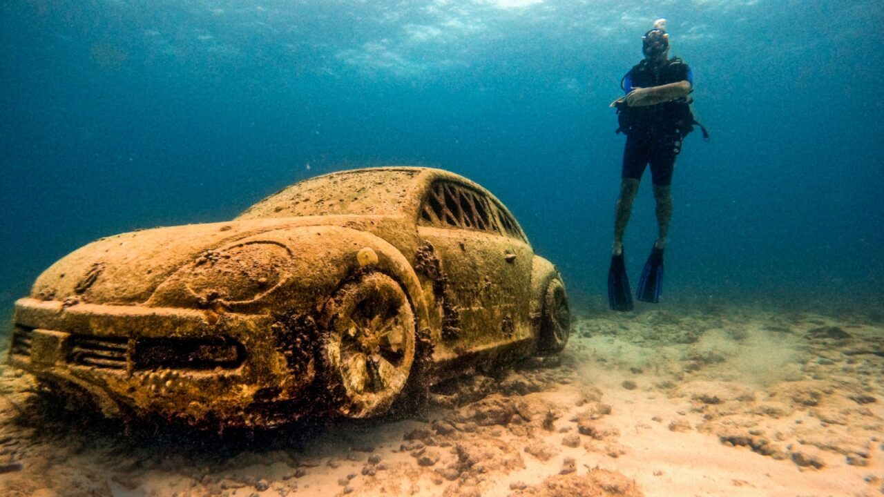 A diver explores a sandy ocean floor where a car covered in marine growth rests. The scene shows underwater tranquility with clear blue water and a mysterious sunken vehicle
