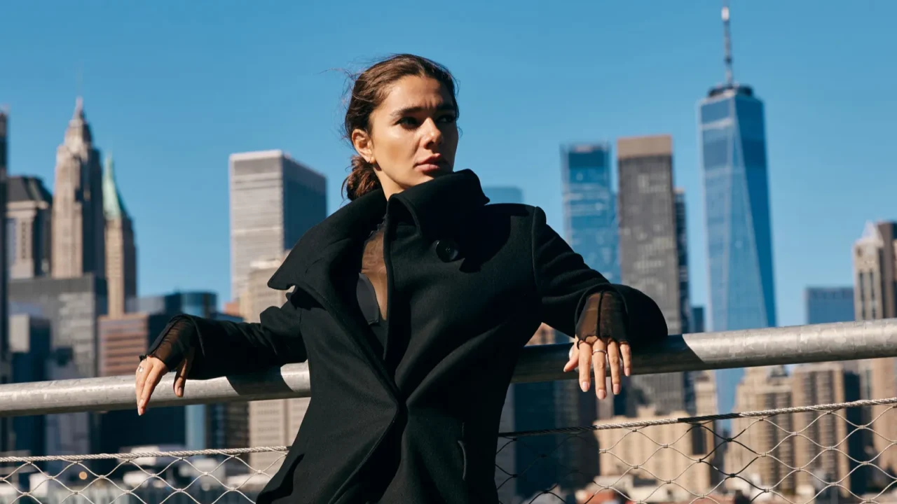 a woman in a black coat poses against the new