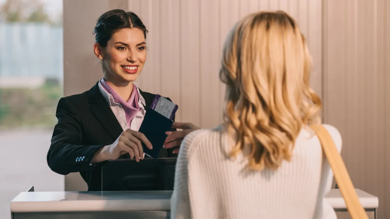 Smiling worker giving passport with boarding pass to young woman at check-in desk in airport.