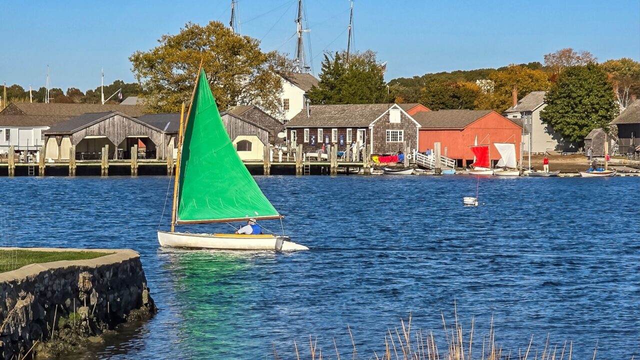 Mystic, Connecticut USA October 19, 2024 Sunny afternoon view of sailboat beyond a charming stone seawall and golden seagrass overlooking the Mystic Seaport on the Mystic River.