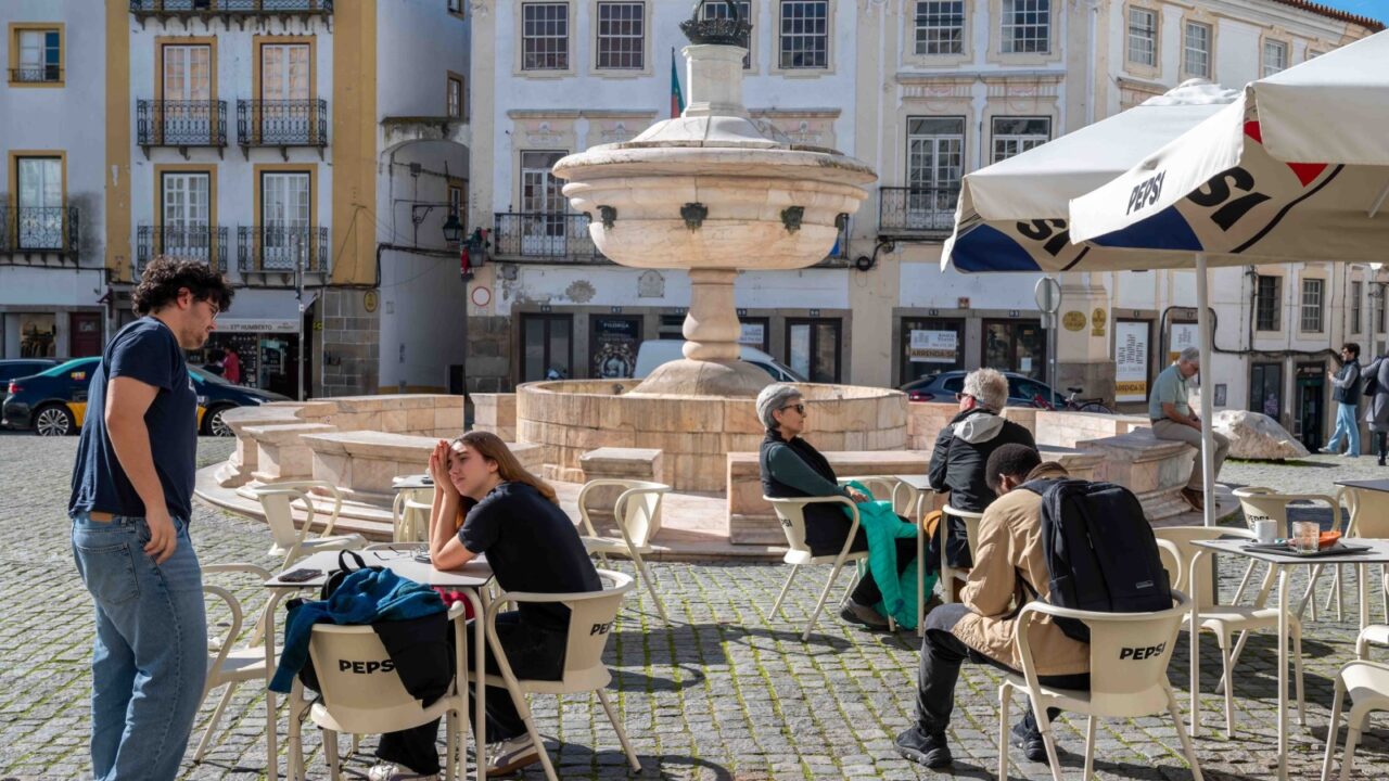 Evora, Portugal, 11-19-2024, People in outdoor restaurant tables with shade umbrellas on the cobblestone town square Praca do Giraldo beside an ancient fountain with quaint stores and buildings in the