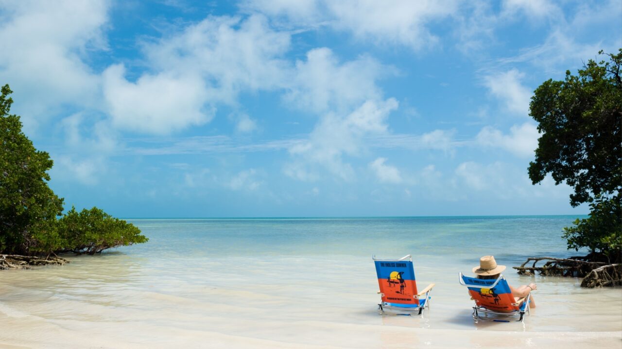 Person relaxing in beach chair beside empty chair on calm tropical ocean shoreline with blue sky and copy space