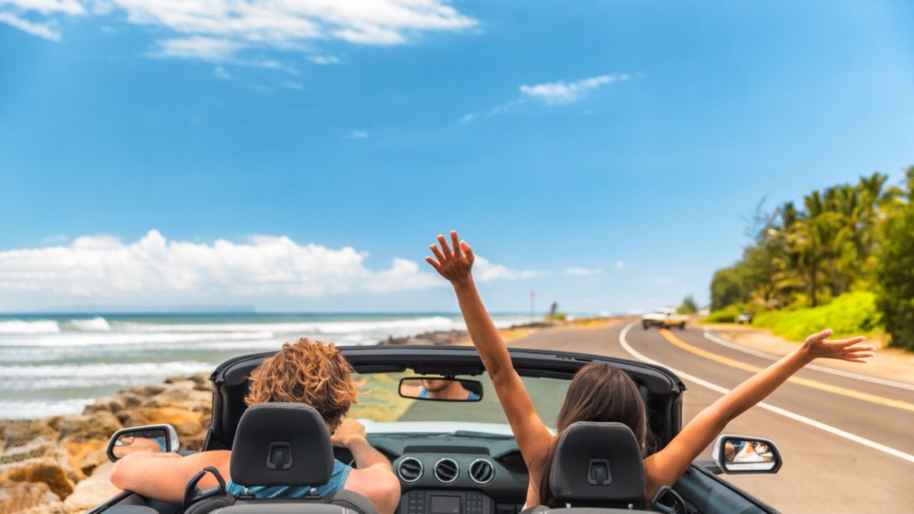 Road trip car holiday happy couple driving convertible car on summer travel Hawaii vacation. Woman with arms up having fun, young man driver