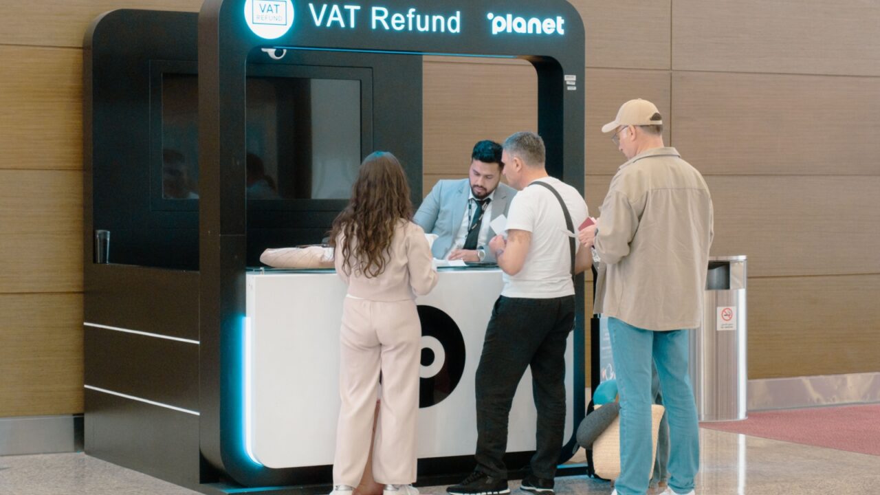 People standing at a VAT refund kiosk at an airport, interacting with a staff member. The group appears to be engaged in a financial transaction. Dubai, UAE - 06-04-2025