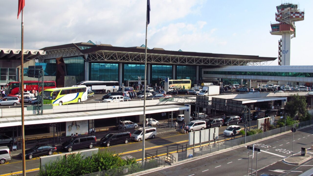 Airport terminal exterior with traffic, buses, cars, glass architecture, checkered control tower, bustling traveler activity.