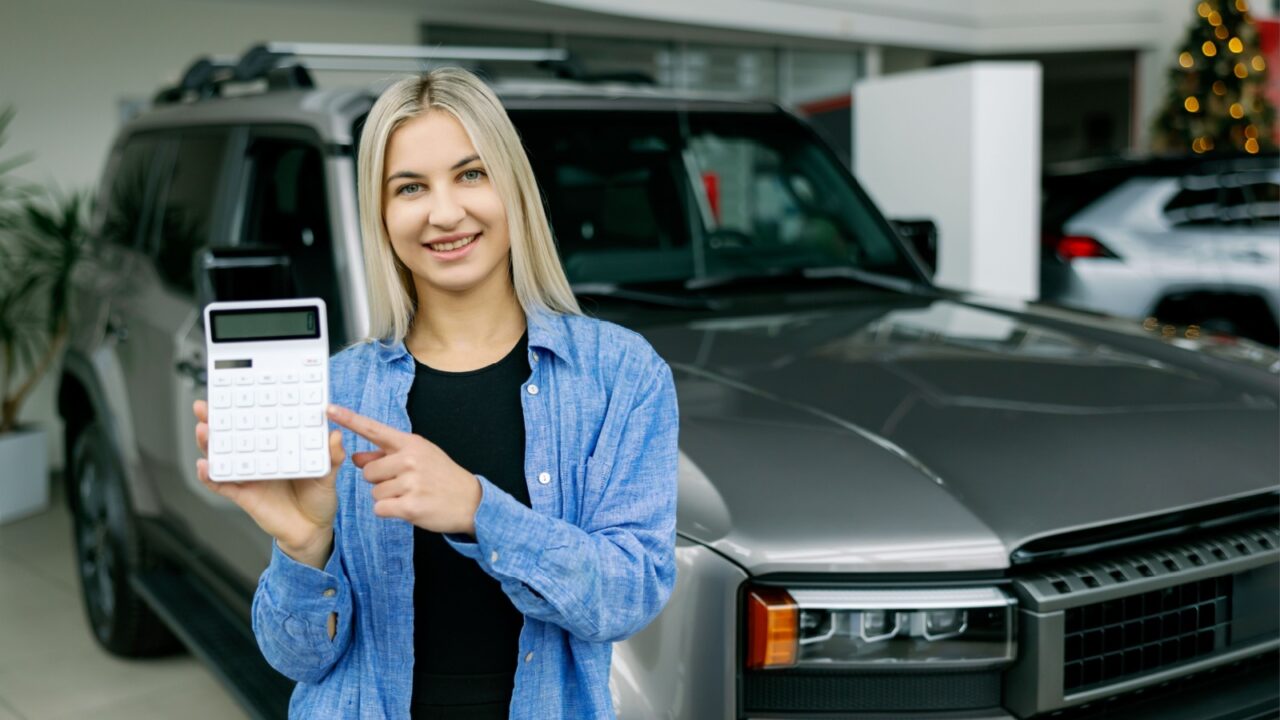 Smiling saleswoman pointing at calculator while calculating car price for customer in dealership