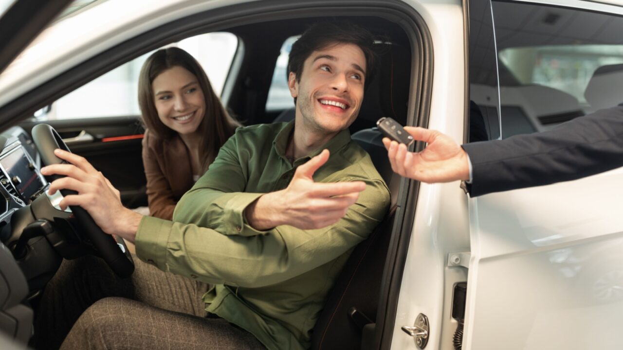 Happy millennial couple taking car key from auto salesman, sitting inside modern automobile at dealership, panorama. Cheery young family buying new vehicle at modern showroom
