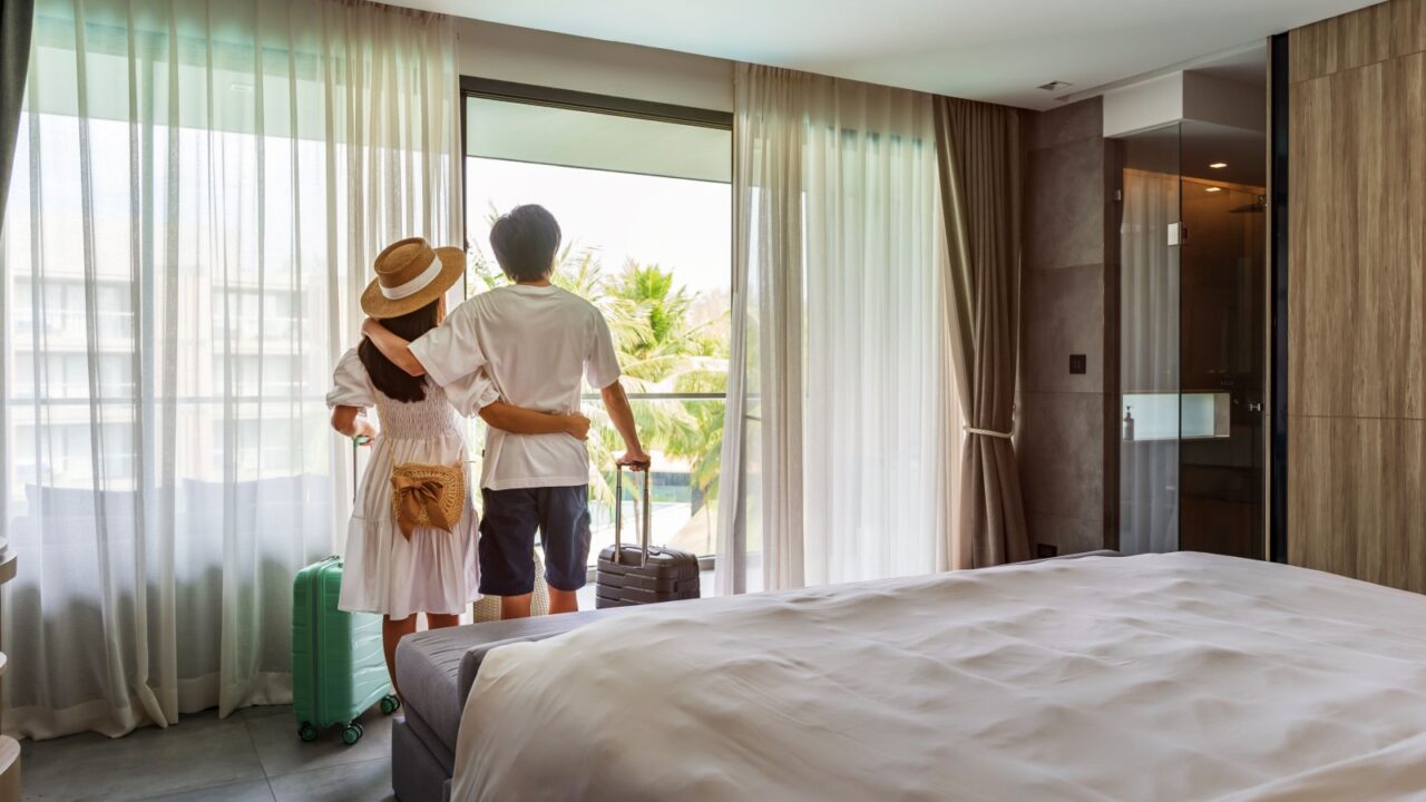 Young couple traveler opening the curtains and looking at the view from the window of a hotel room while on summer vacation, Travel lifestyle concept