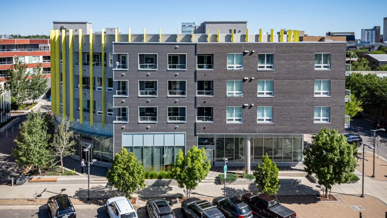 Large building with a yellow trim sits in front of a parking lot. The parking lot is full of cars, including a black car and a white car. The building has a modern design and he is a hotel