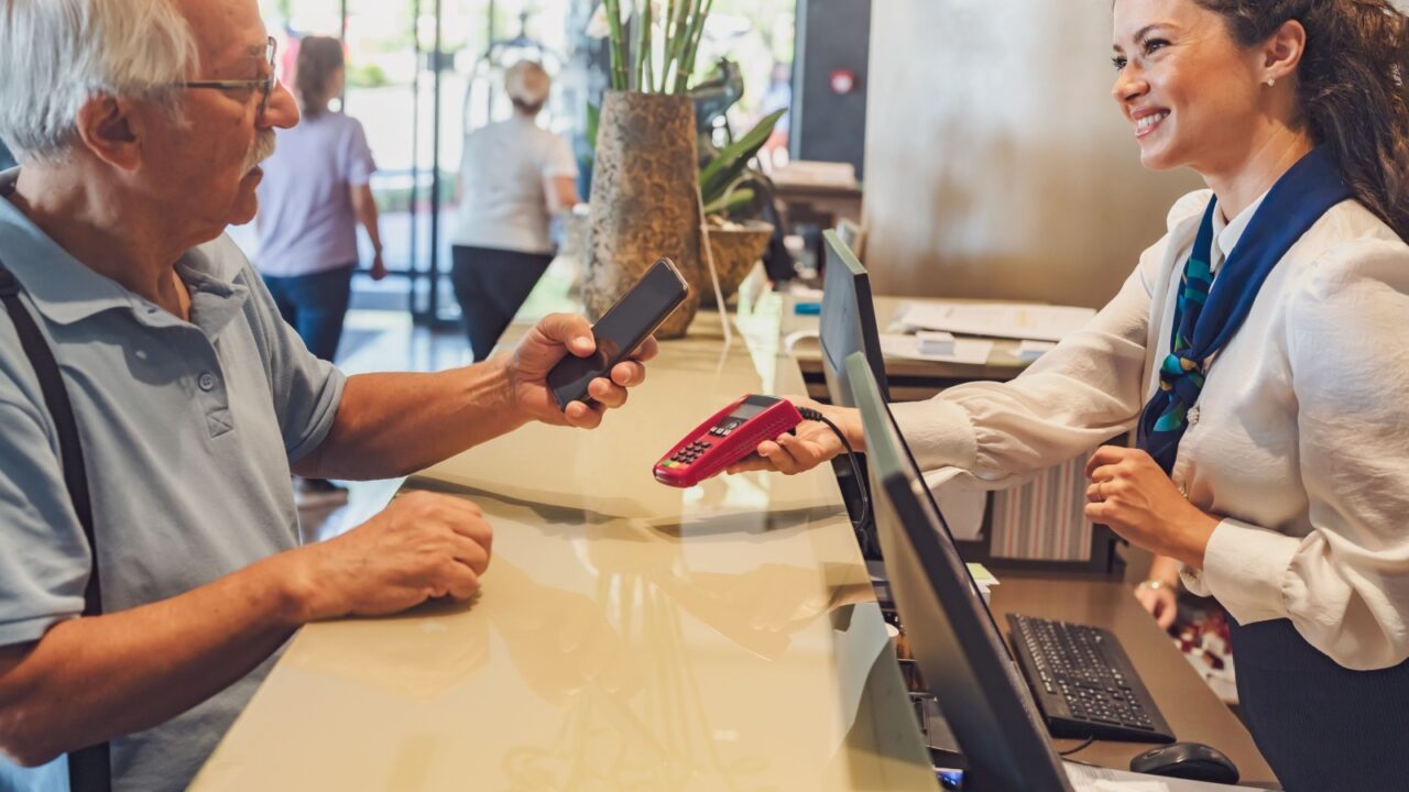 A senior couple at the hotel front desk makes contactless payments with a smartphone. Seniors traveling and using new technologies