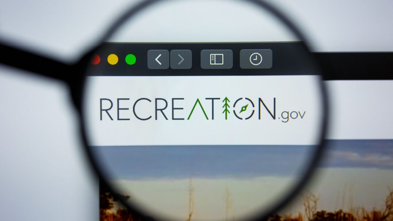 Magnifying glass highlights RECREATION.gov logo, tent, tree, and compass symbols on computer screen.