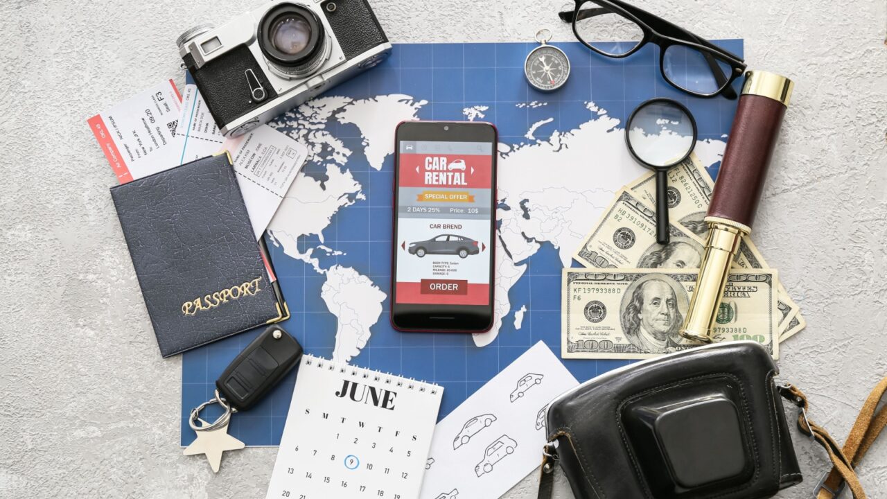 Mobile phone with open car rent app, map, passport, photo camera and money on light background