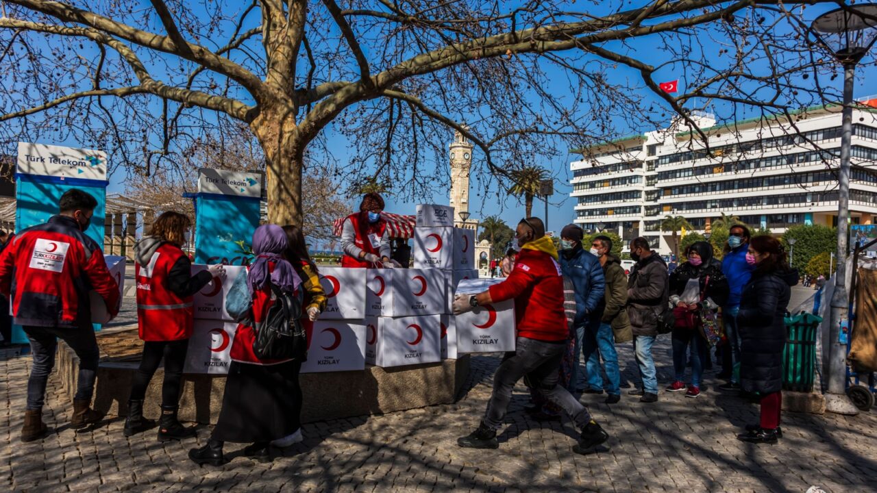 Volunteers in red vests distribute Kızılay aid boxes to people, Turkish flag and clock tower visible.