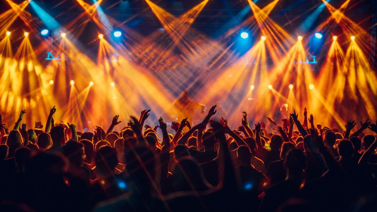 Open air music festival with crowd in foreground having fun, dancing and rising their hands. A performer is singing on stage during the concert while silhouettes of people having fun at concert.