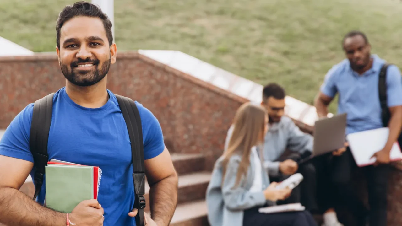 a confident indian student stands holding books on campus steps