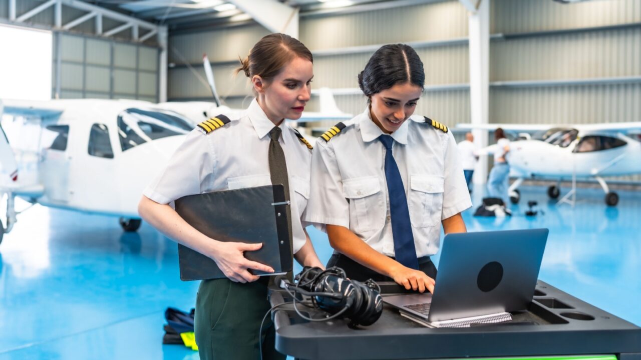 Two smiling female pilots in uniform working on a laptop inside a hangar, collaborating next to an airplane, showcasing their aviation expertise