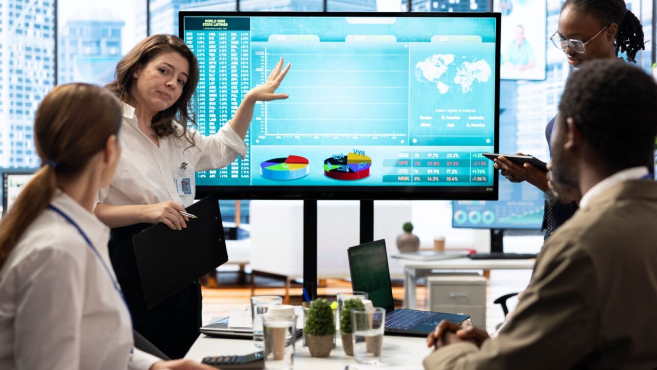 Business associates collaborating at the conference table in boardroom, utilizing smart technology and monthly statistics to present a detailed analysis of market trends and economic forecasts.