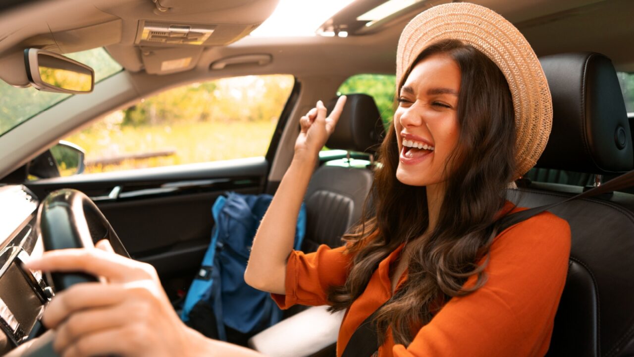 Overjoyed woman tourist enjoying car ride alone, listening to music, singing songs and dancing while driving auto with backpack nearby on passenger seat