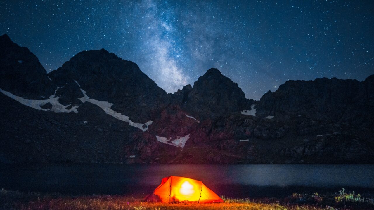 A glowing tent under the Milky Way in a high-altitude mountain landscape. Perfect for concepts like solitude, outdoor adventure, stargazing, and freedom.
