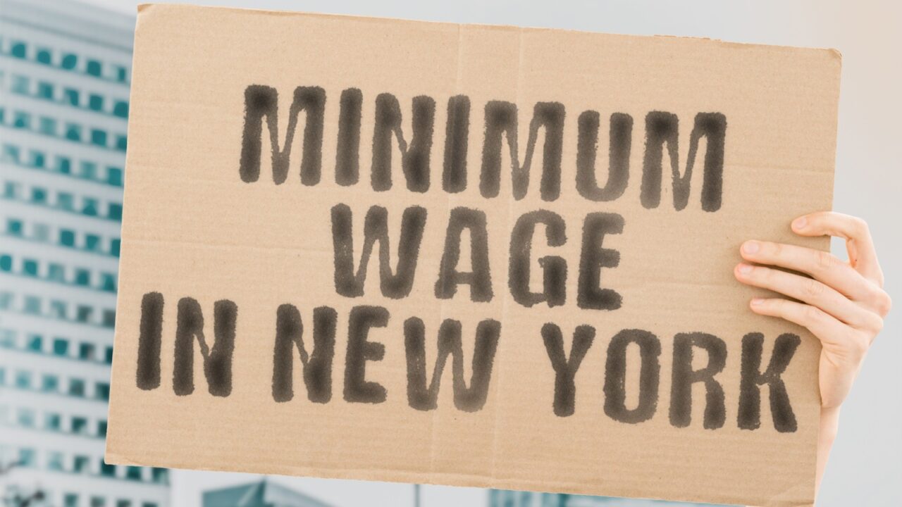 The question " Minimum wage in New York " on a banner in men's hand with blurred background. Business. Money. Revenue. Earnings. Payment. Job. Occupation