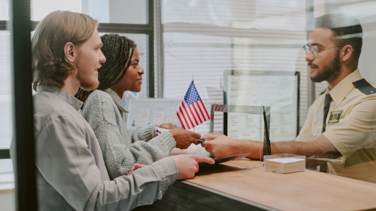 Diverse couple handing passports at immigration office counter with officer verifying documents and flag in background. Multi-ethnic group interacting in official setting