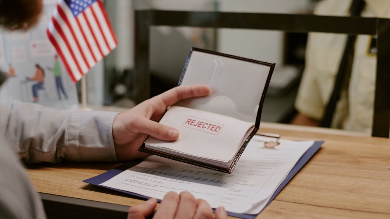 Hand holding passport stamped with rejected over desk in immigration office, with American flag in background. Person interacting with immigration officer behind desk