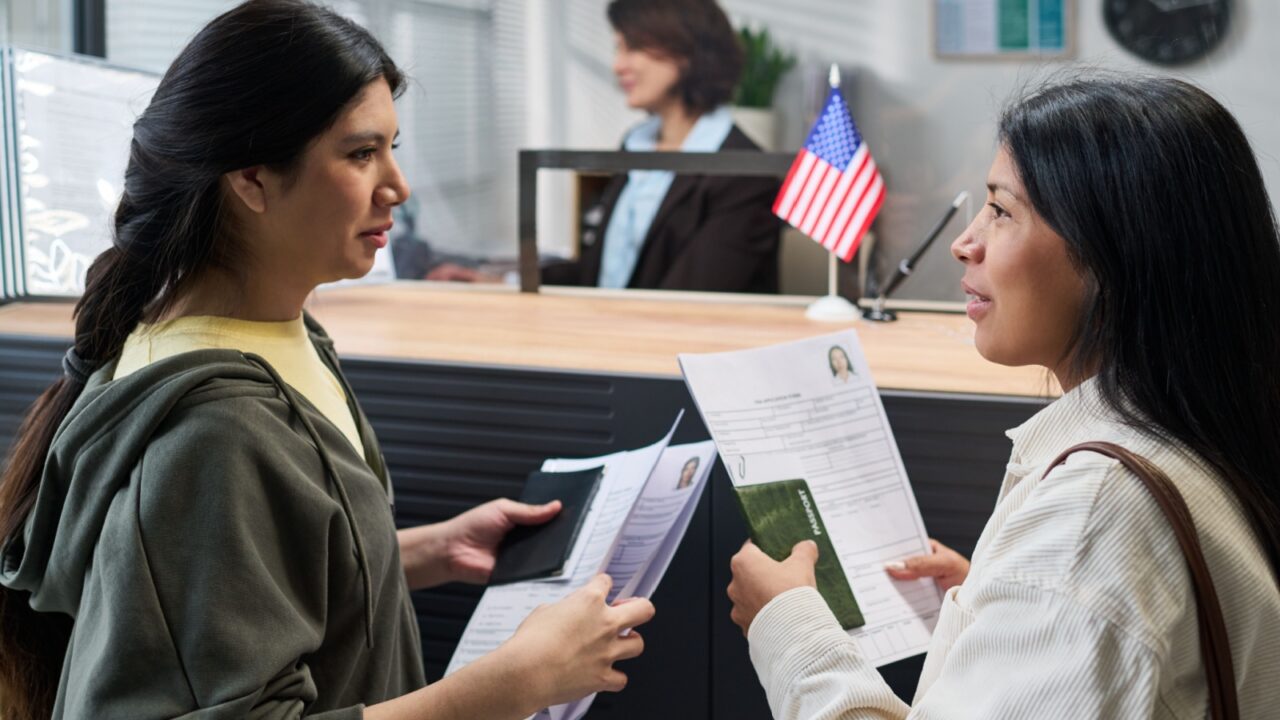 Two young adult Hispanic women standing at visa center counter holding documents and passports, engaging in conversation while waiting for service, female staff member visible in background