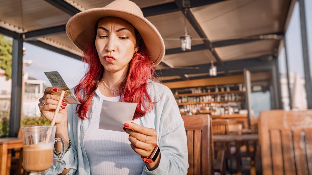 Young woman sitting at a restaurant table with a displeased expression checking the bill and paying in cash