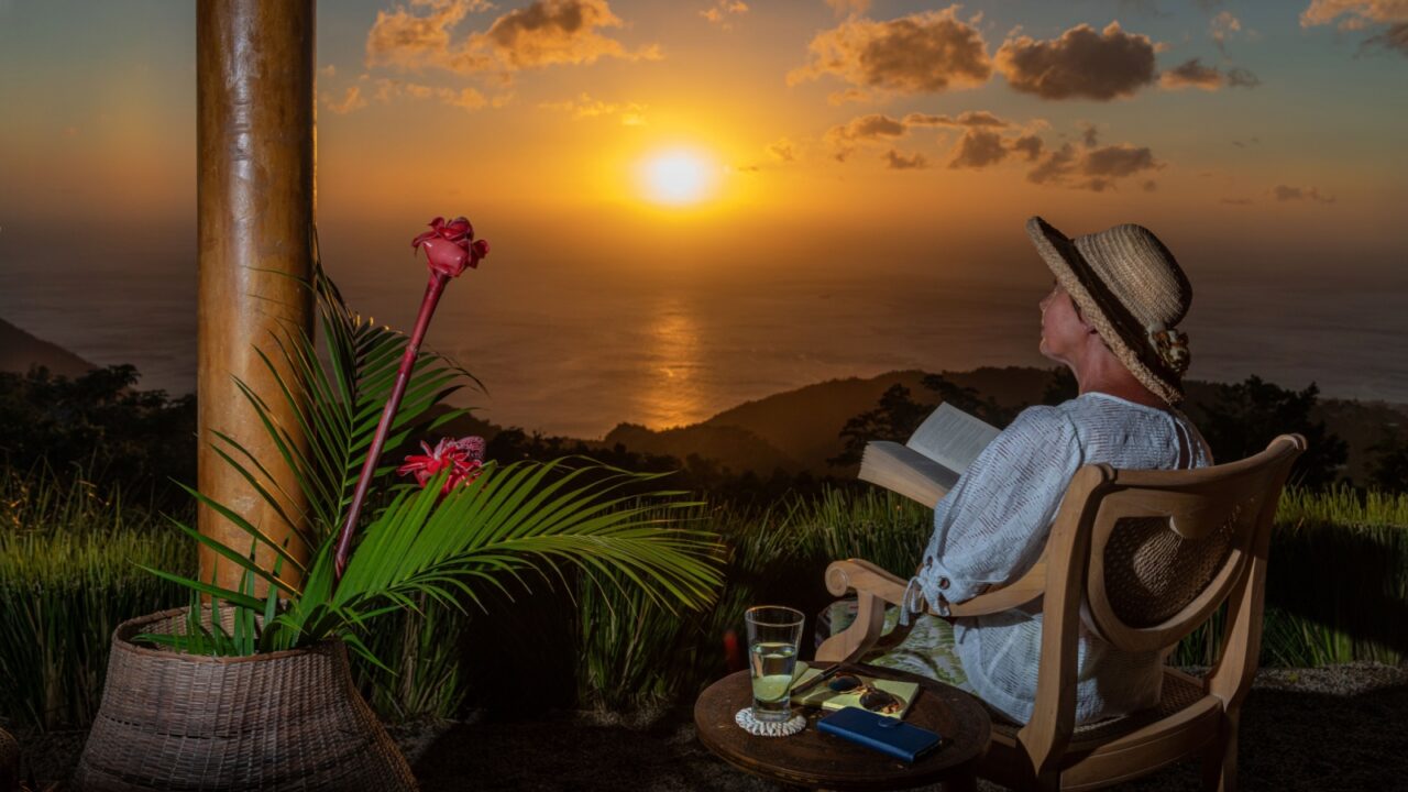 Woman in a summer hat enjoys reading,working,relaxing at beautiful Caribbean sunset. Patio with large red torch ginger lily flowers. Digital nomads, remote workers.