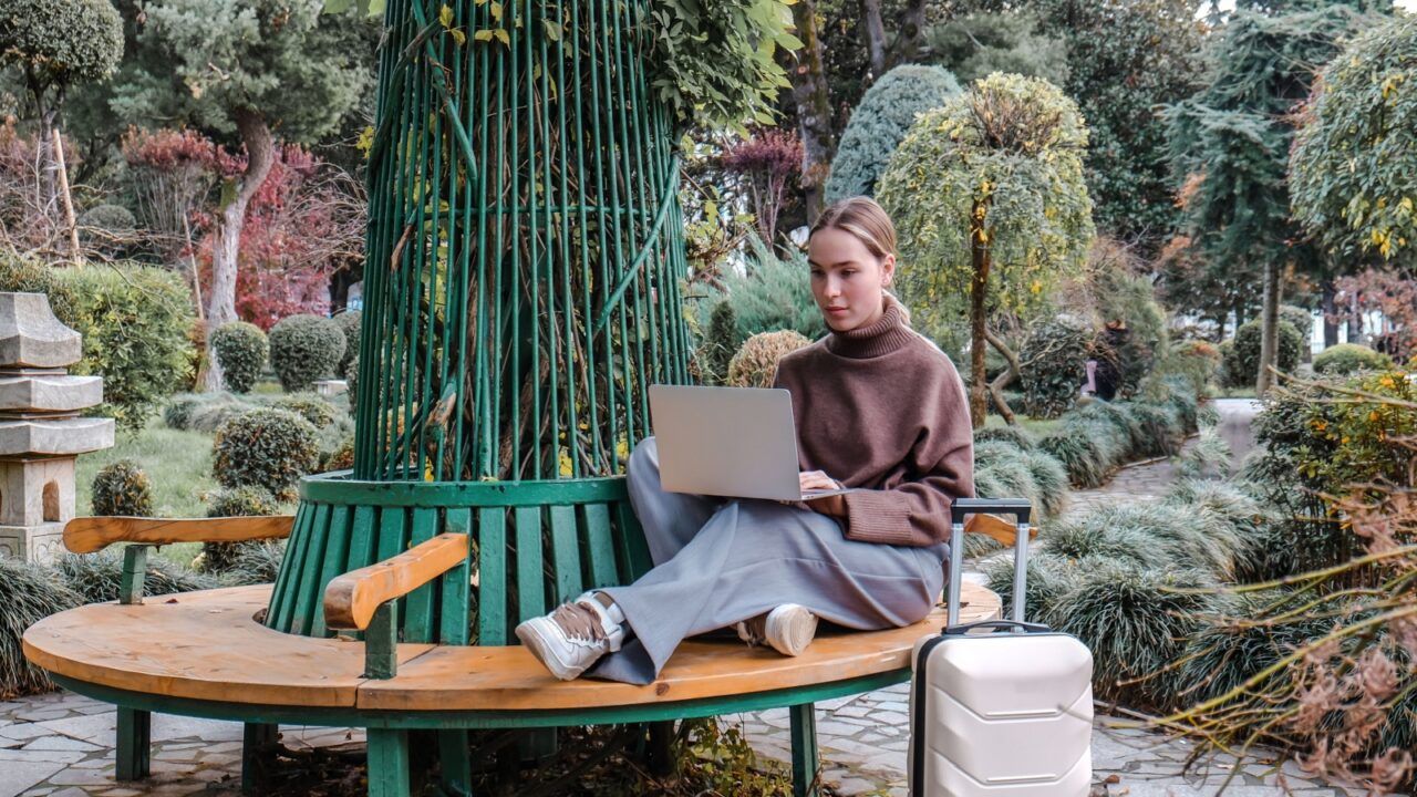 Person sitting on circular wooden bench in garden, using laptop beside suitcase, surrounded by tranquil greenery.