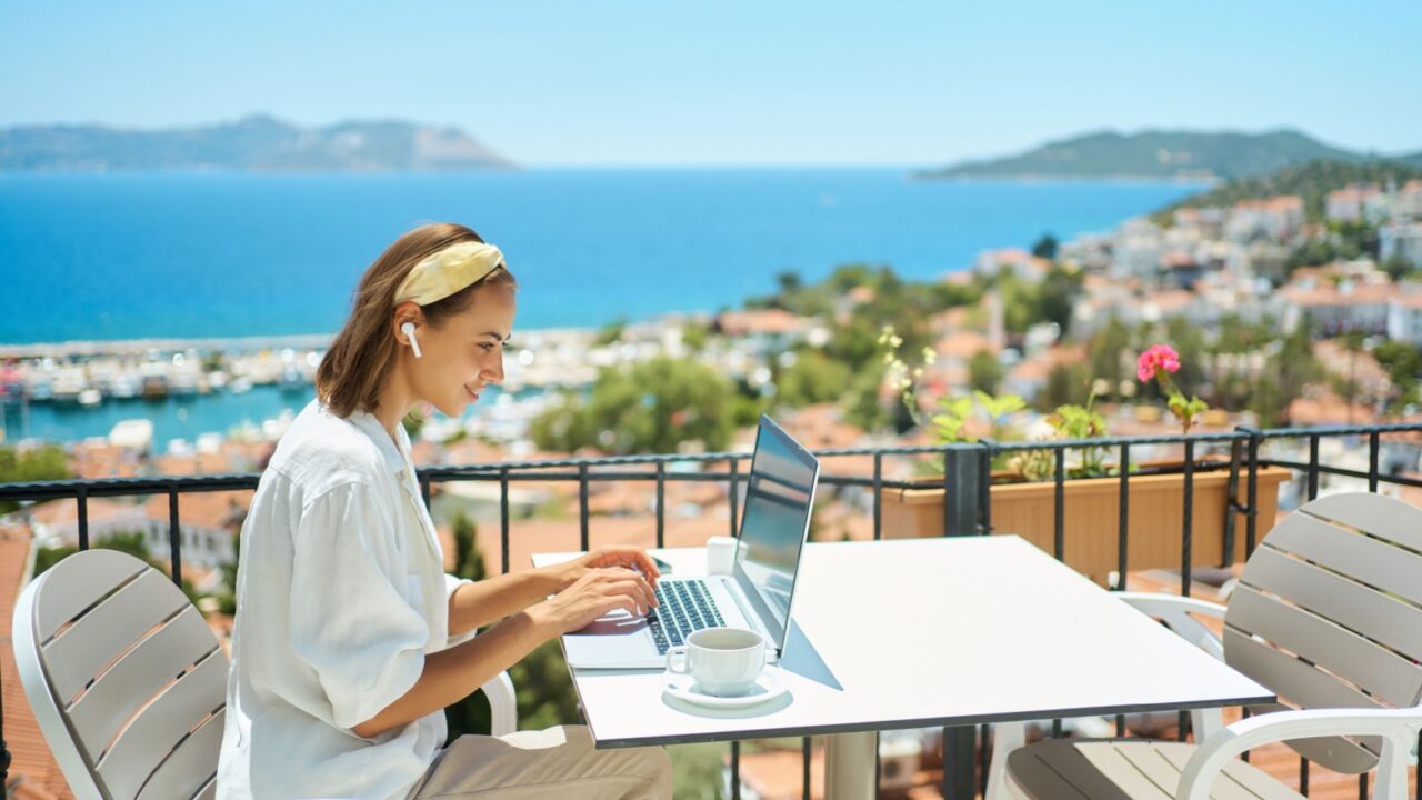 Young woman using laptop computer at cafe balcony of resort hotel with sea view, working typing emails browsing online enjoying drinking coffee