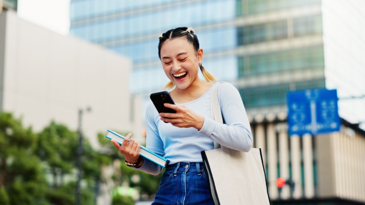 Woman, happy and phone in city for social media, good news and student loan approval in Tokyo. Japanese girl, mobile and excited in town for scholarship opportunity, achievement and academic report