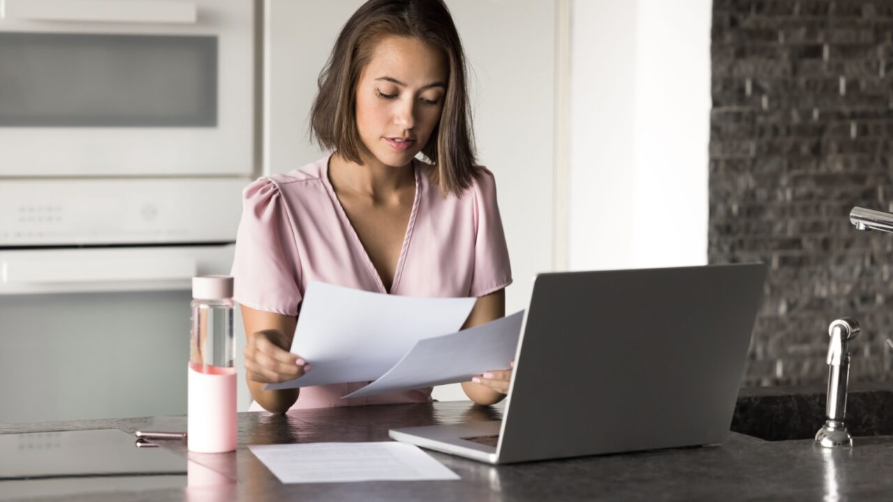 Young 25s woman sits at kitchen counter with laptop, focused on reviewing important documents, working on task, reviewing reports, contracts, or assignments. Teleworking, freelance, budget management