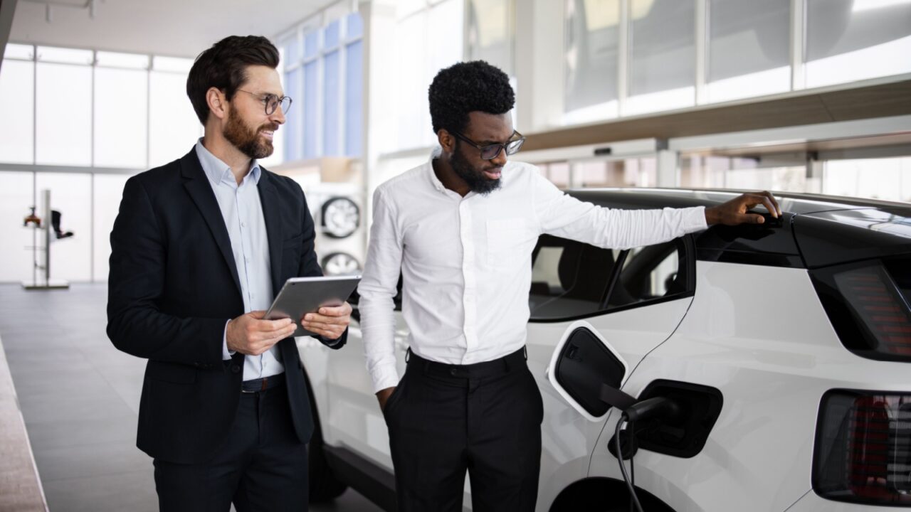Dealer sells electric SUV to businessman at modern car dealership. Professional consultation with use of tablet. Display of car features, benefits of electric vehicle.