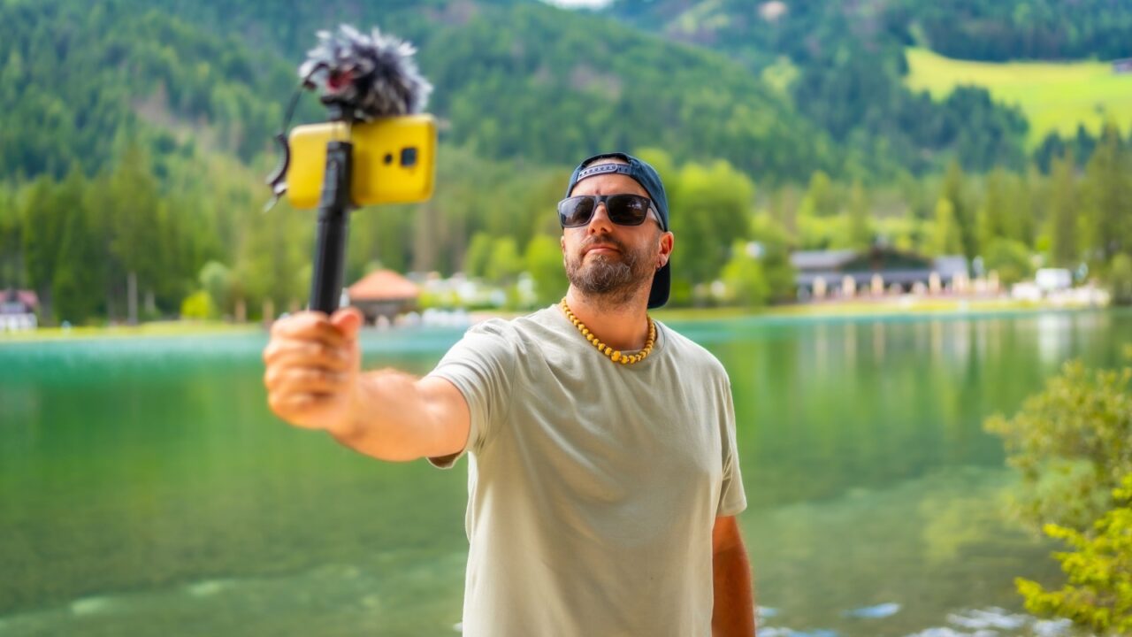 Content creator filming a video with a smartphone and external microphone at lake dobbiaco in the italian dolomites during summer