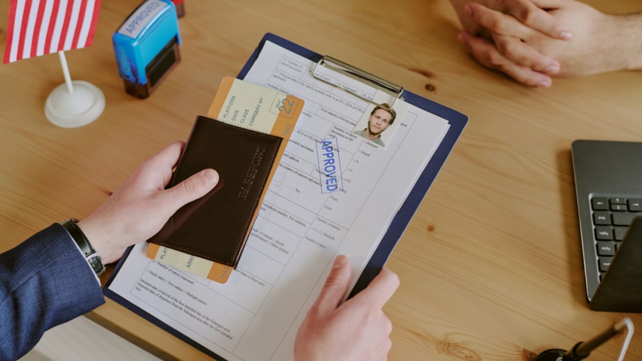 Handing passport and documents across table during visa interview. American flag on table alongside laptop, envelope, and clipboard with approved form visible